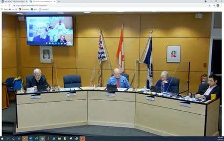 Screenshot of the council meeting chamber showing members at the dais and a remote participant on a wall-mounted screen