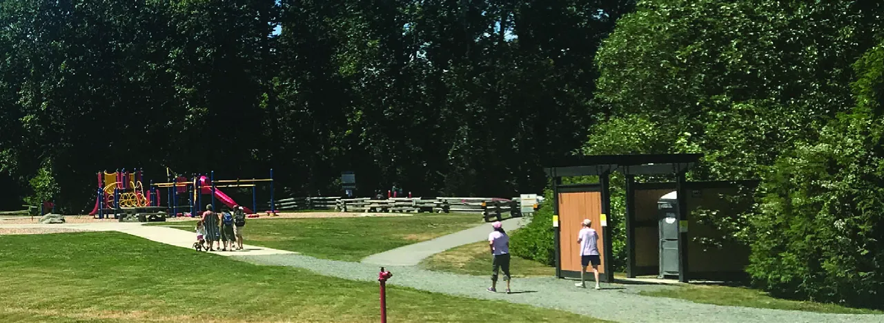 A sunny park scene with a playground on the left and a gravel walking path where people are walking toward a wooden structure