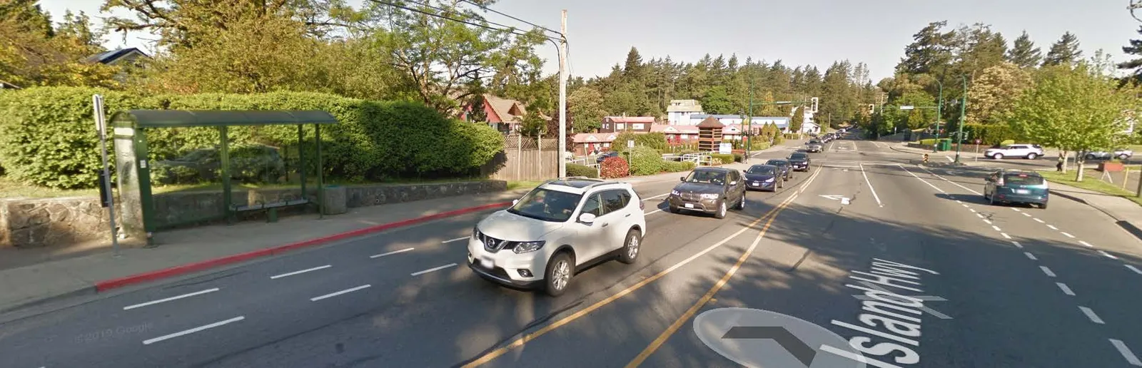 Street-level view of Island Highway showing a transit stop, vehicle traffic, and surrounding greenery