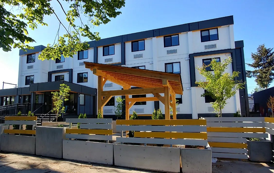 Exterior view of Aneki Housing for Women, showing a modern modular building with a wooden gazebo and fencing in the foreground