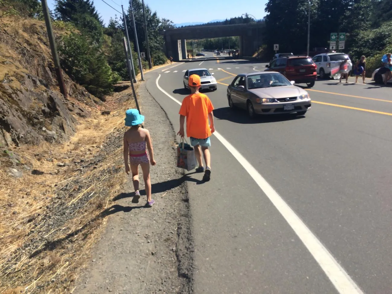 Two children walking along a narrow, unpaved shoulder of a busy road with cars passing nearby and an overpass visible in the background