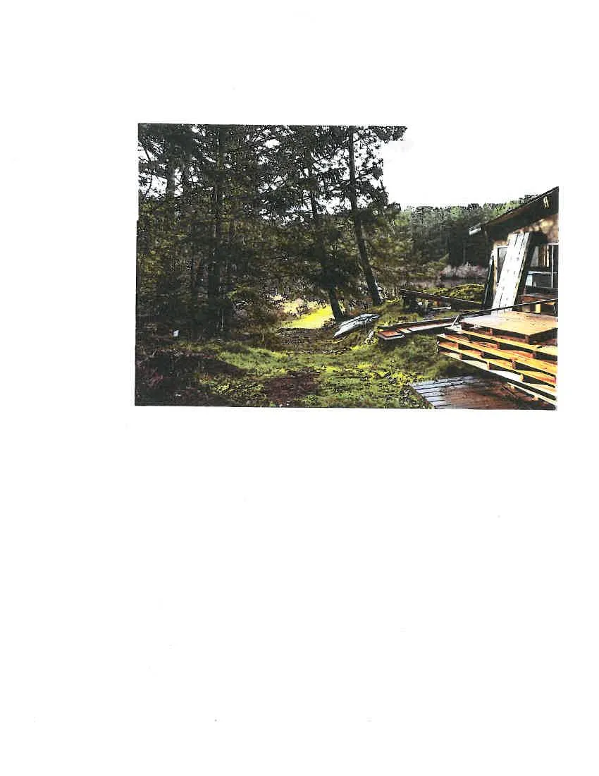 A photograph showing construction materials, including stacked lumber and pallets, next to a building with a view through trees toward a body of water.
