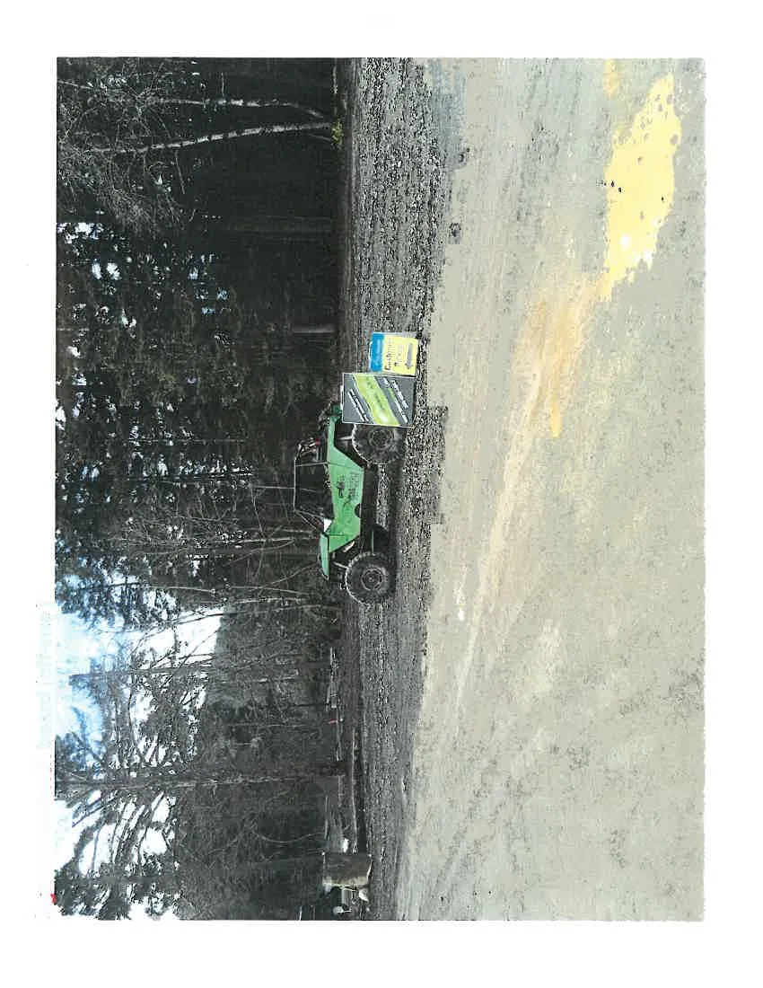 A green off-road vehicle parked next to a sandwich board sign in a gravel area bordered by trees.