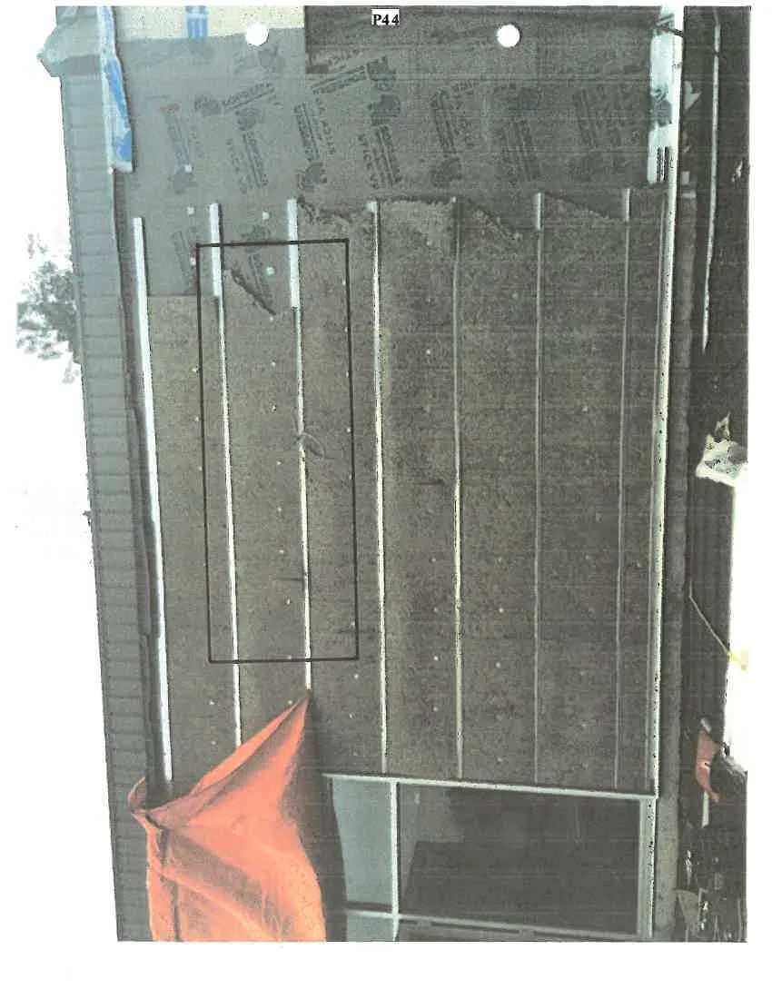 Photograph of a building exterior wall during construction or repair. The wall assembly includes grey insulation panels, vertical furring strips, and a SOPREMA STICK VP moisture barrier. A black rectangular box is drawn around a section of the insulation boards on the left side of the wall. An orange tarp is visible at the bottom of the frame.